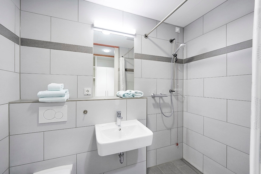 Rechts ist die Dusche. Direkt daneben ist das Waschbecken mit einem Spiegel. Links ist die Toilette. Auf der Ablage liegen aufgefaltete Handtücher.